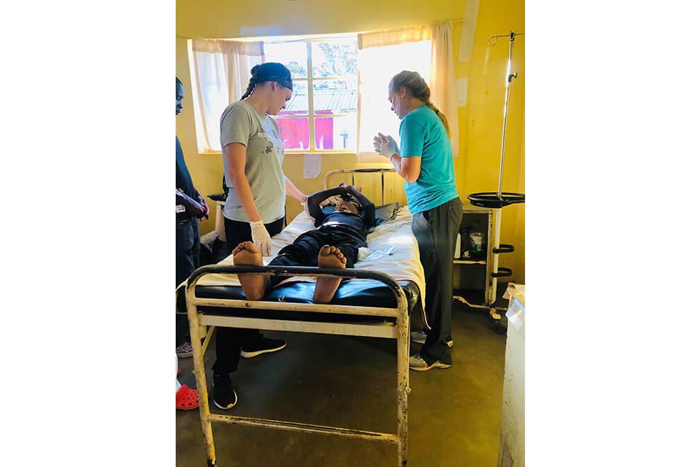 Two female physical therapy students stand on either side of patient's bed