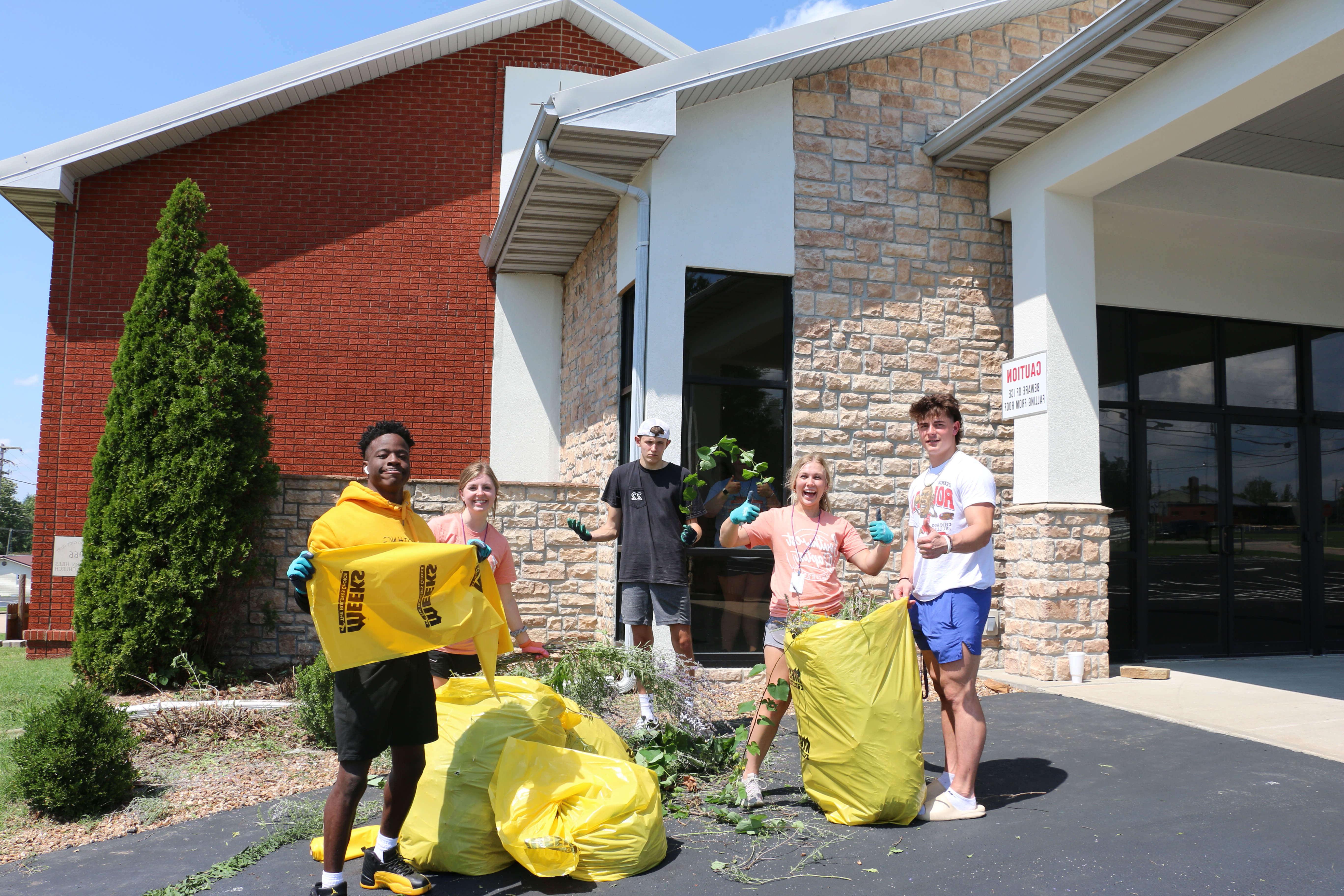 group of students poses for picture holding yellow bags and working on pulling weeds outside church building