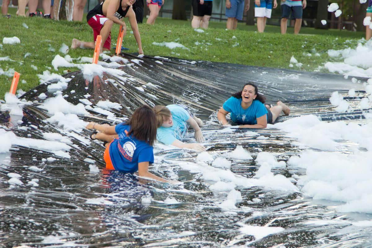 two female students laugh while sliding down the slip and slide