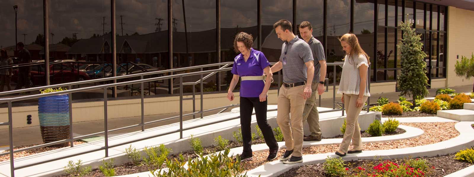 two students walk behind instructor who is helping patient walk through physical therapy garden
